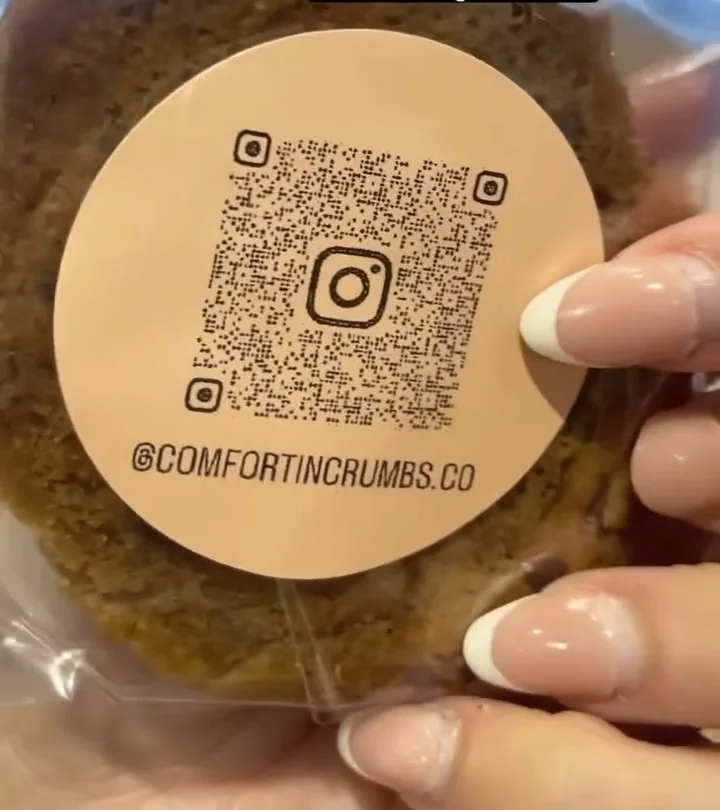 An individually wrapped cookie sealed with a Comfort In Crumbs sticker showing the Instagram QR code and @comfortincrumbs.co